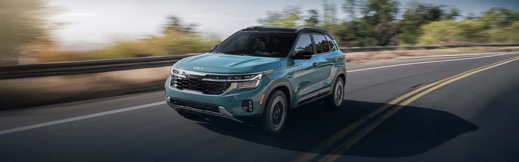 2026 Kia Seltos compact SUV driving on an open highway with a sleek turquoise exterior and scenic roadside landscape in the background.
